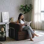 A woman sits barefoot in a brown corduroy armchair reading a magazine in a bright, modern room with a gray concrete wall, white curtains, and a large green plant.
