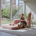 A woman sits on a pink cushioned floor sofa in a bright room, painting on an easel. A small dog rests on the sofa beside a small wooden side table with a mug.