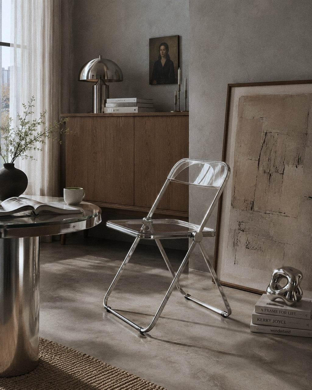 A clear acrylic folding chair with a silver metal frame, styled in a modern, minimalist room with concrete floors and neutral-toned furniture.