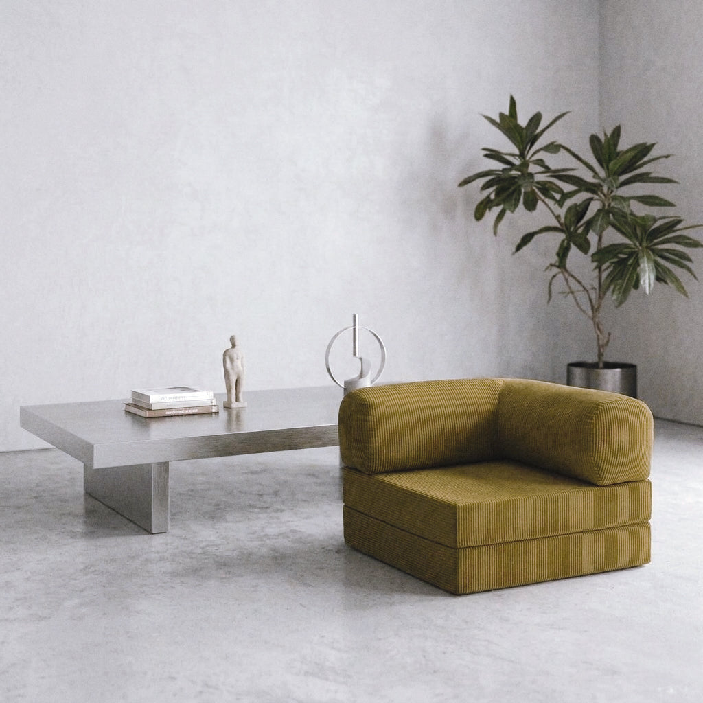 An olive green corduroy armchair in a modern minimalist room with a concrete floor and textured gray walls, next to a low concrete coffee table and a potted plant.
