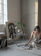 A woman in a beige slip dress sits on a woven rug reading a book in a minimalist concrete room, next to a clear metal folding chair holding a glass vase with green leaves.