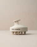 A round, textured off-white hair massager brush with a handle, featuring the text 'skin meets' printed in black on the front, set against a minimalist beige background.