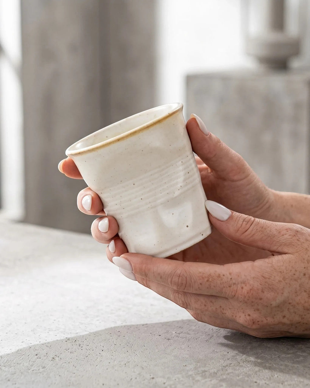 ESTYNE Studio Form Cup in off-white glazed ceramic with a hand-formed silhouette held in hands on a concrete surface.