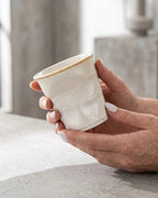 ESTYNE Studio Form Cup in off-white glazed ceramic with a hand-formed silhouette held in hands on a concrete surface.