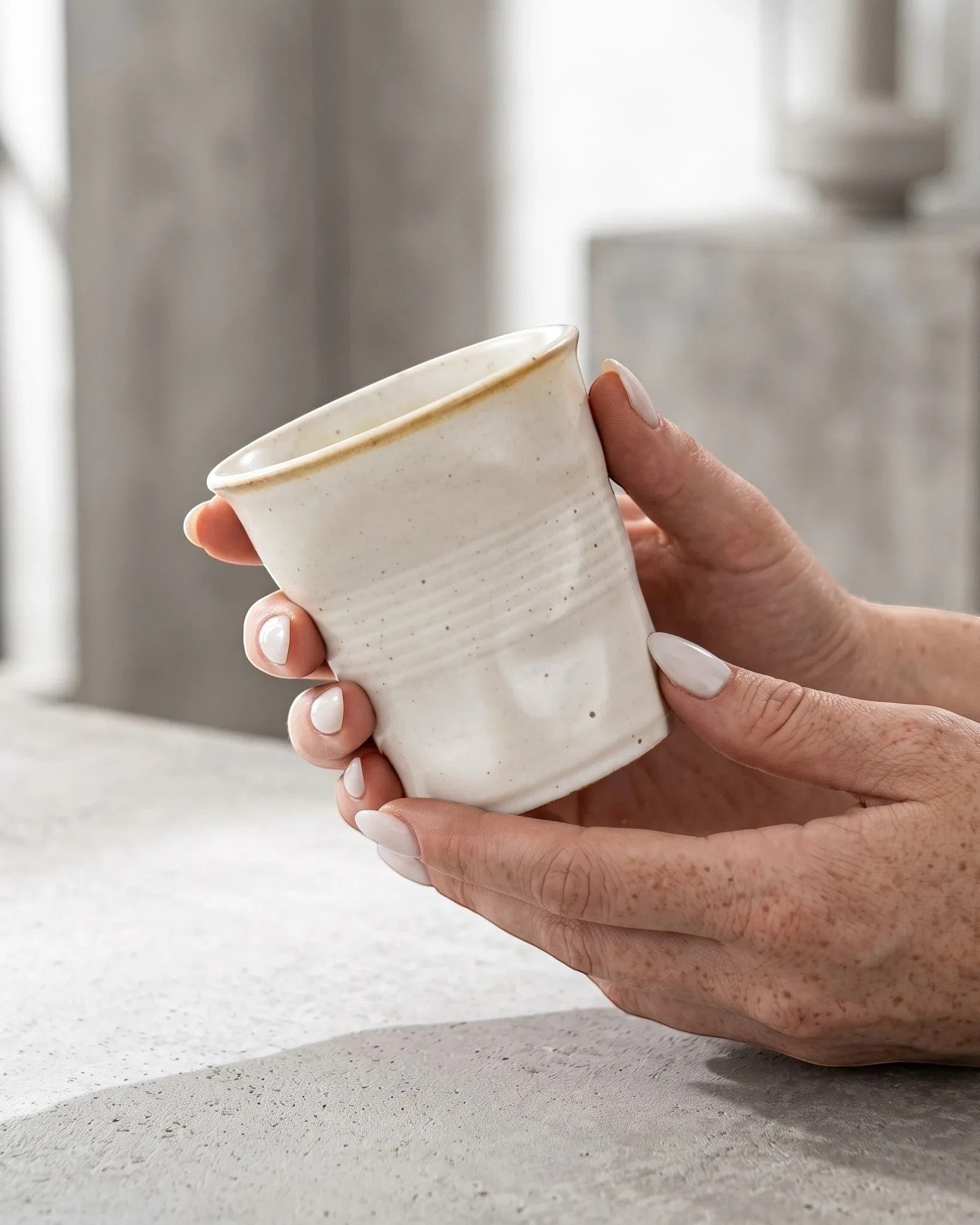 ESTYNE Studio Form Cup in off-white glazed ceramic with a hand-formed silhouette held in hands on a concrete surface.