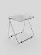 Novage Studios modern folding chair with a transparent acrylic seat and a polished chrome metal frame.